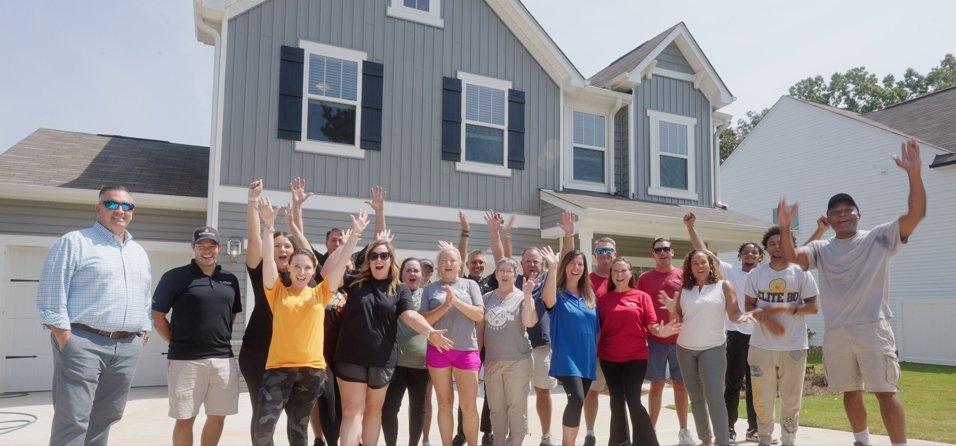 Group of people celebrating in front of a new gray house with arms raised.
