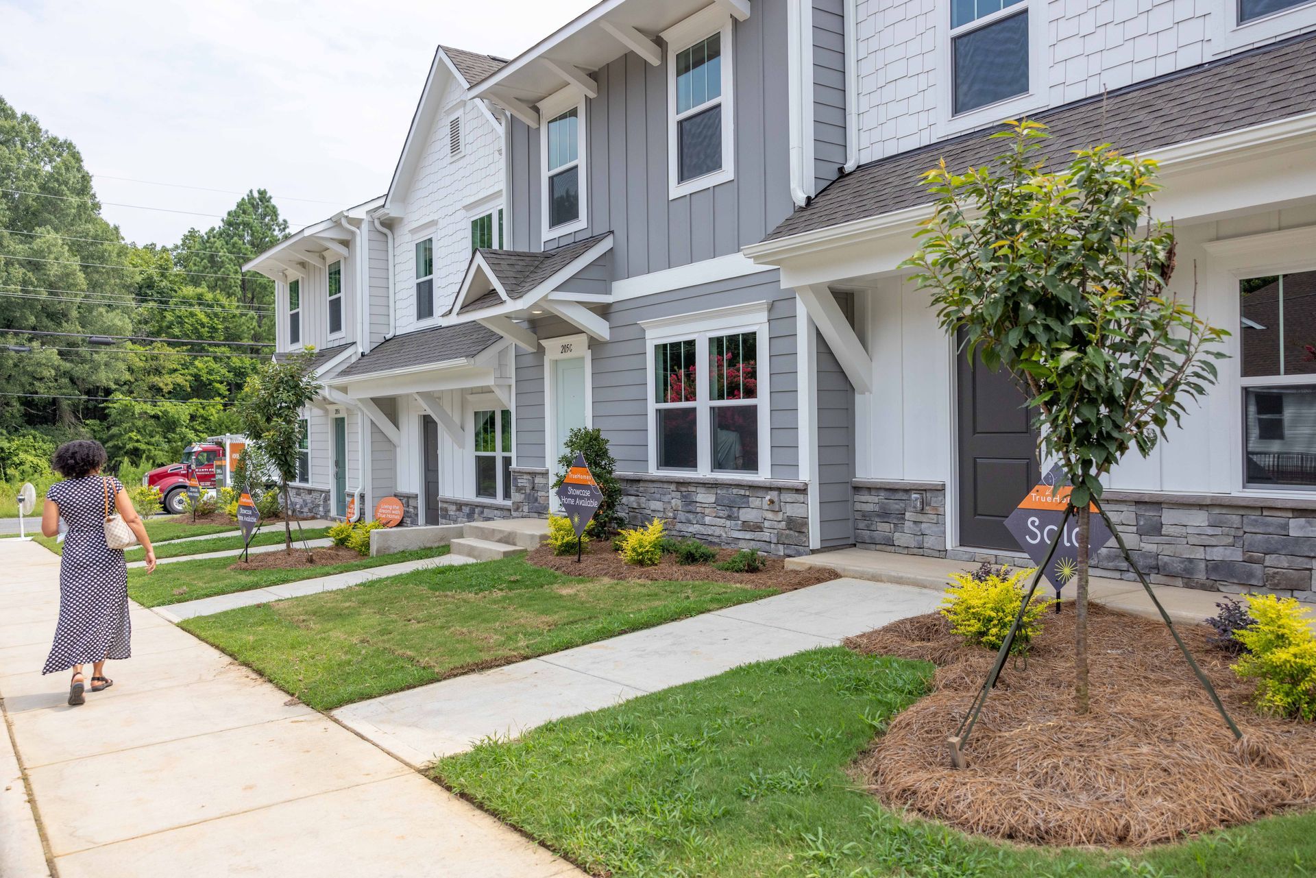 Woman walks toward modern townhomes; gray and white facades with stone accents, sunny day.