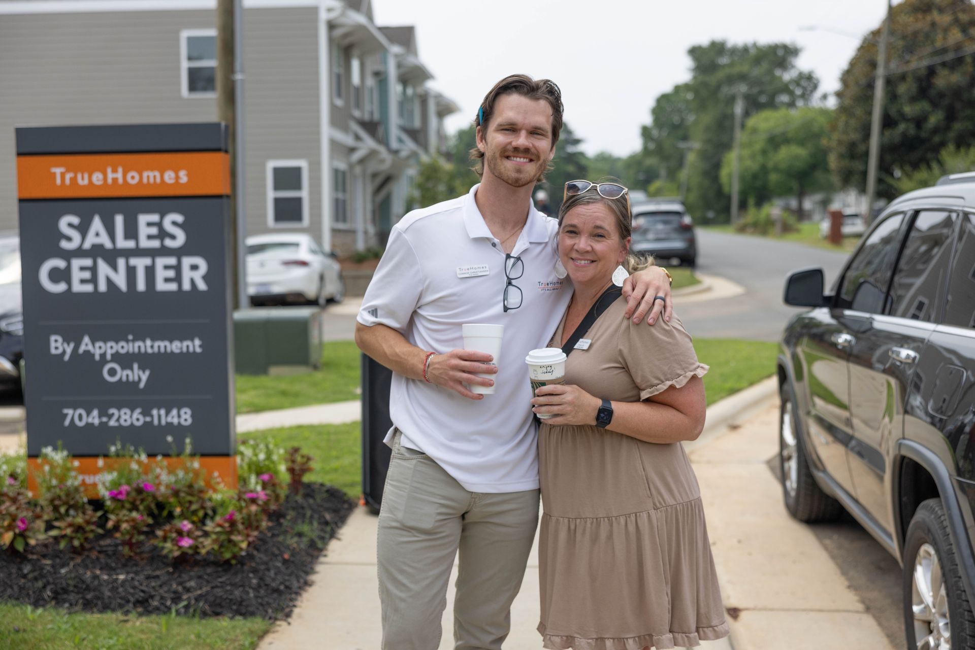 A smiling couple stands by a sales center sign, holding drinks. Buildings and a black SUV are in the background.