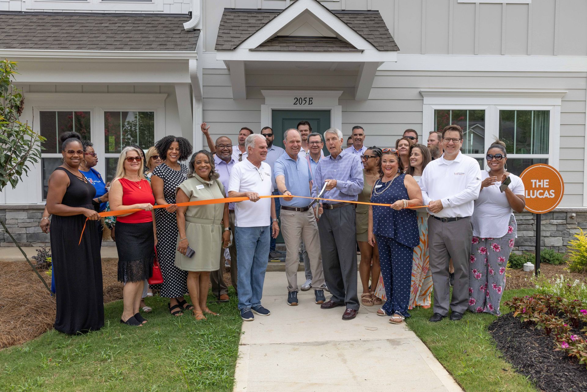 People cutting an orange ribbon in front of a new house, celebrating an opening.