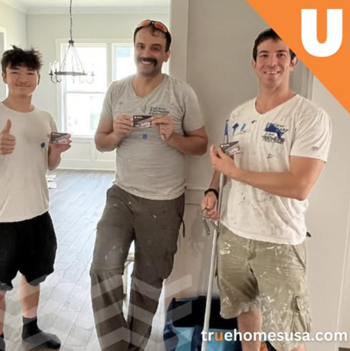 Three men in a house, holding cards, smiling. Paint-splattered clothes, thumbs up.
