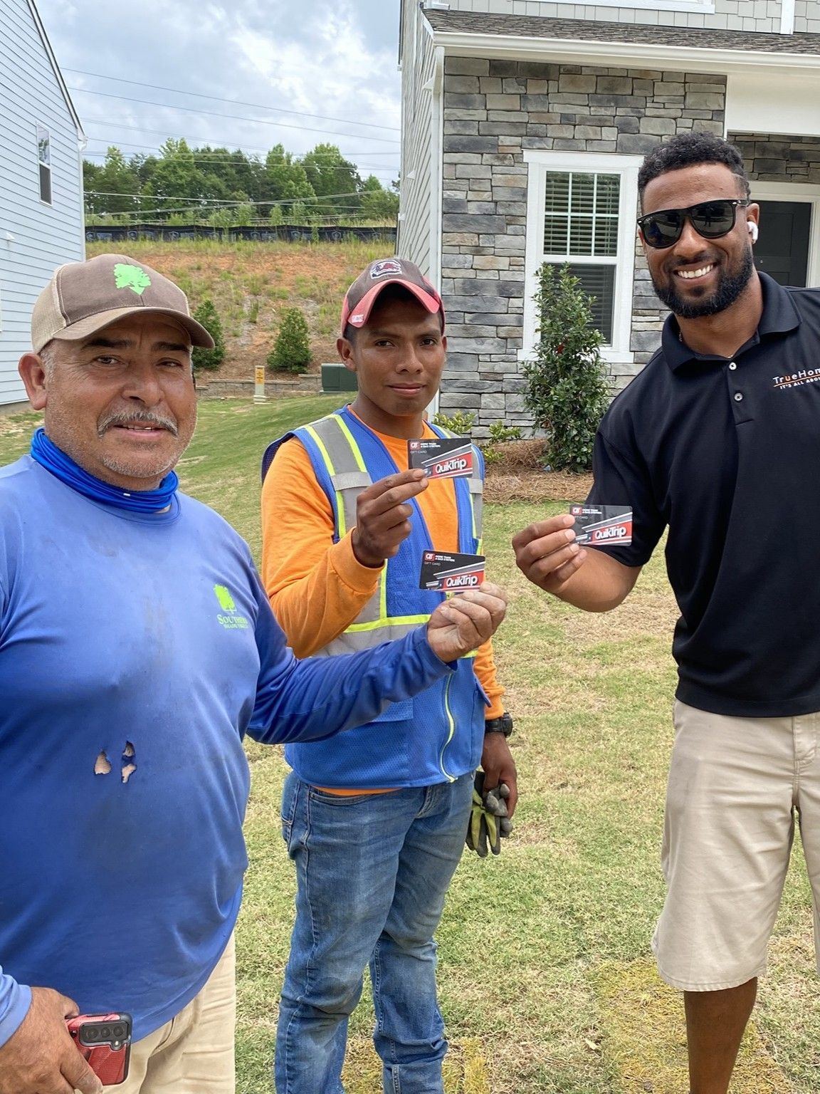 Three men smiling and holding cards. Landscapers in front of a house.