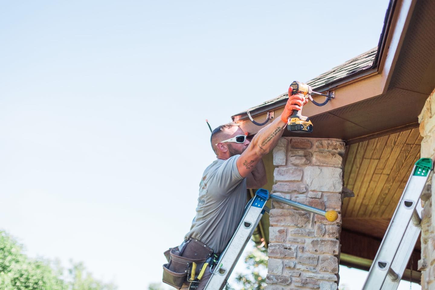 A man is sitting on a ladder fixing a gutter on a house.