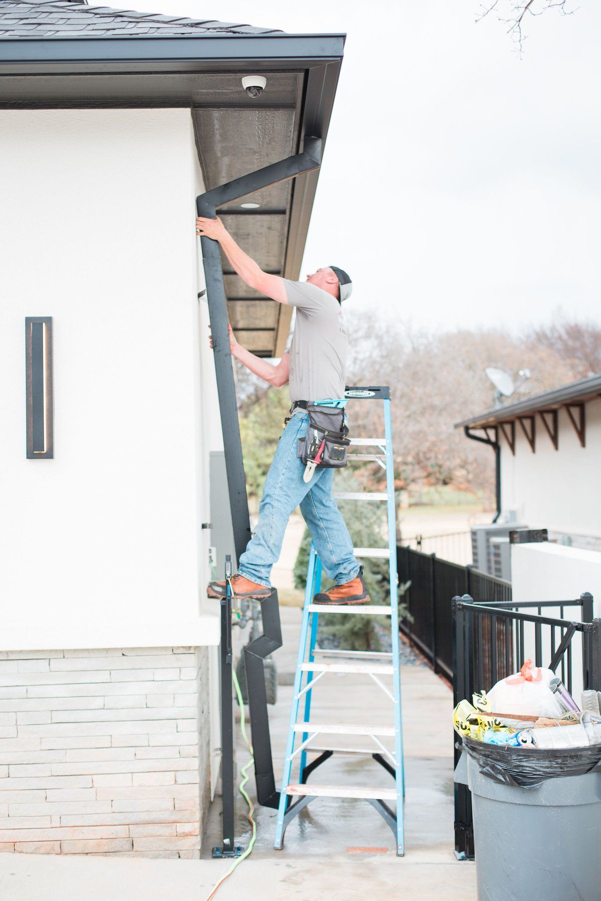 A man is standing on a ladder fixing a gutter on the side of a house.