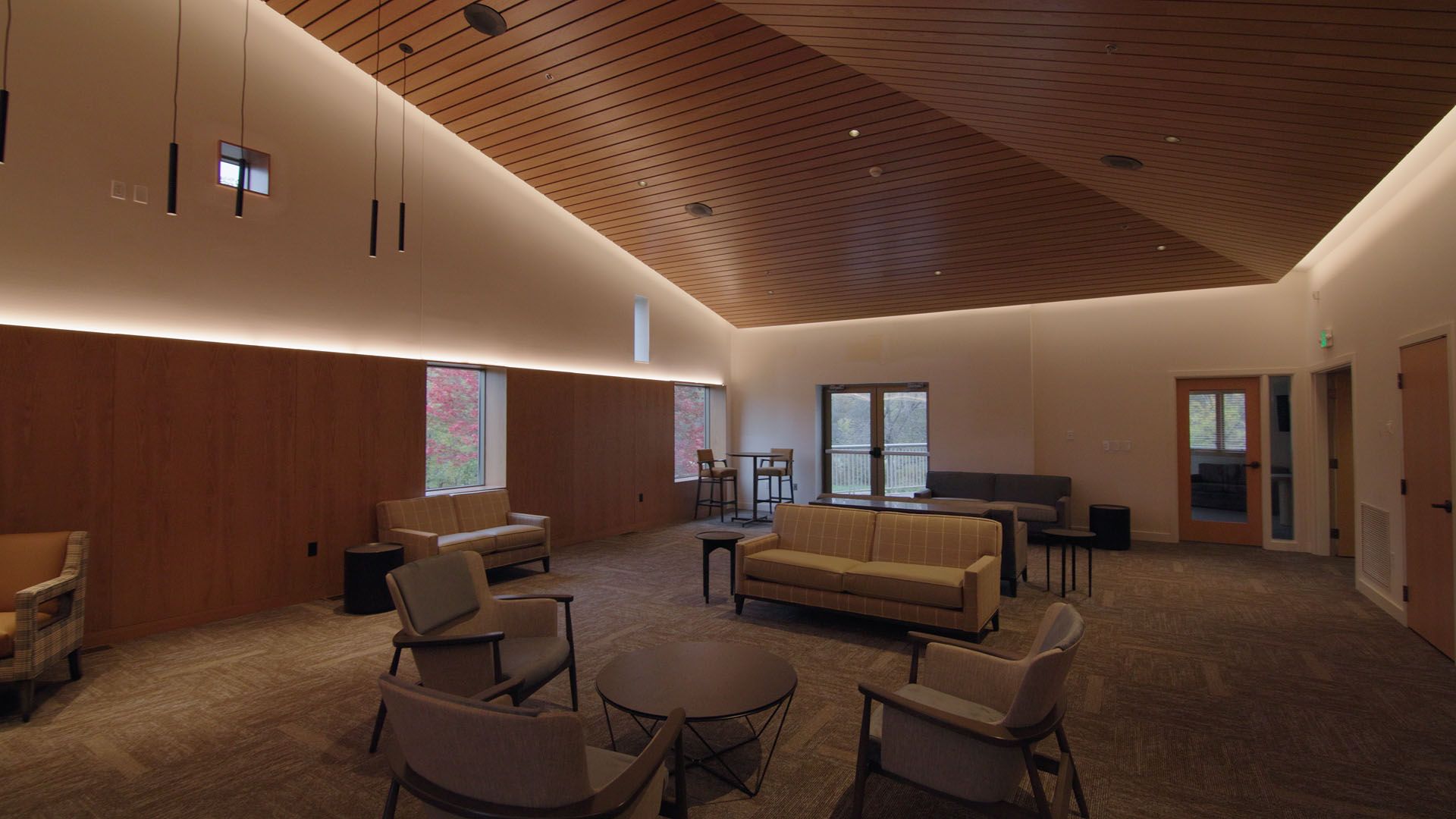 Lounge area with light-colored couches, chairs, and round tables, set against wooden paneling and a wood-paneled ceiling with recessed lighting.