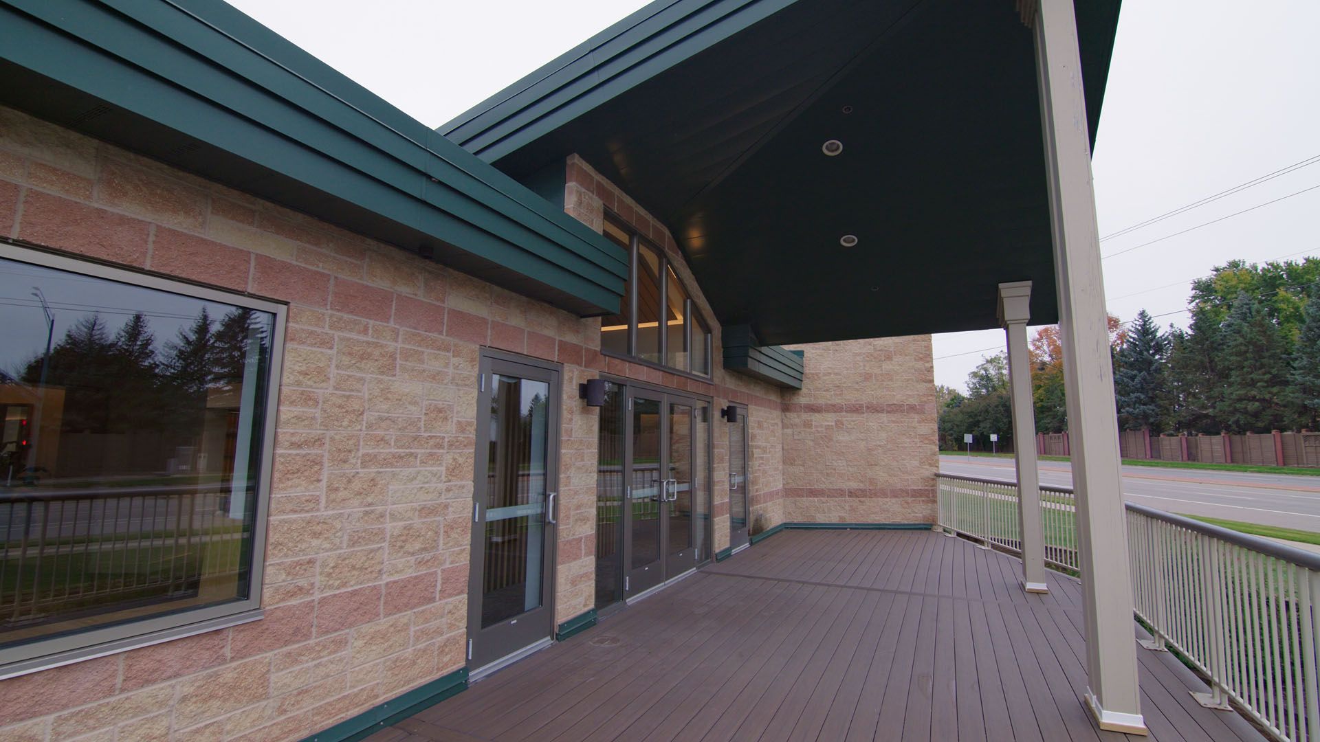 Exterior of a building with brick walls, a dark green roof, and a covered walkway with a wooden deck.