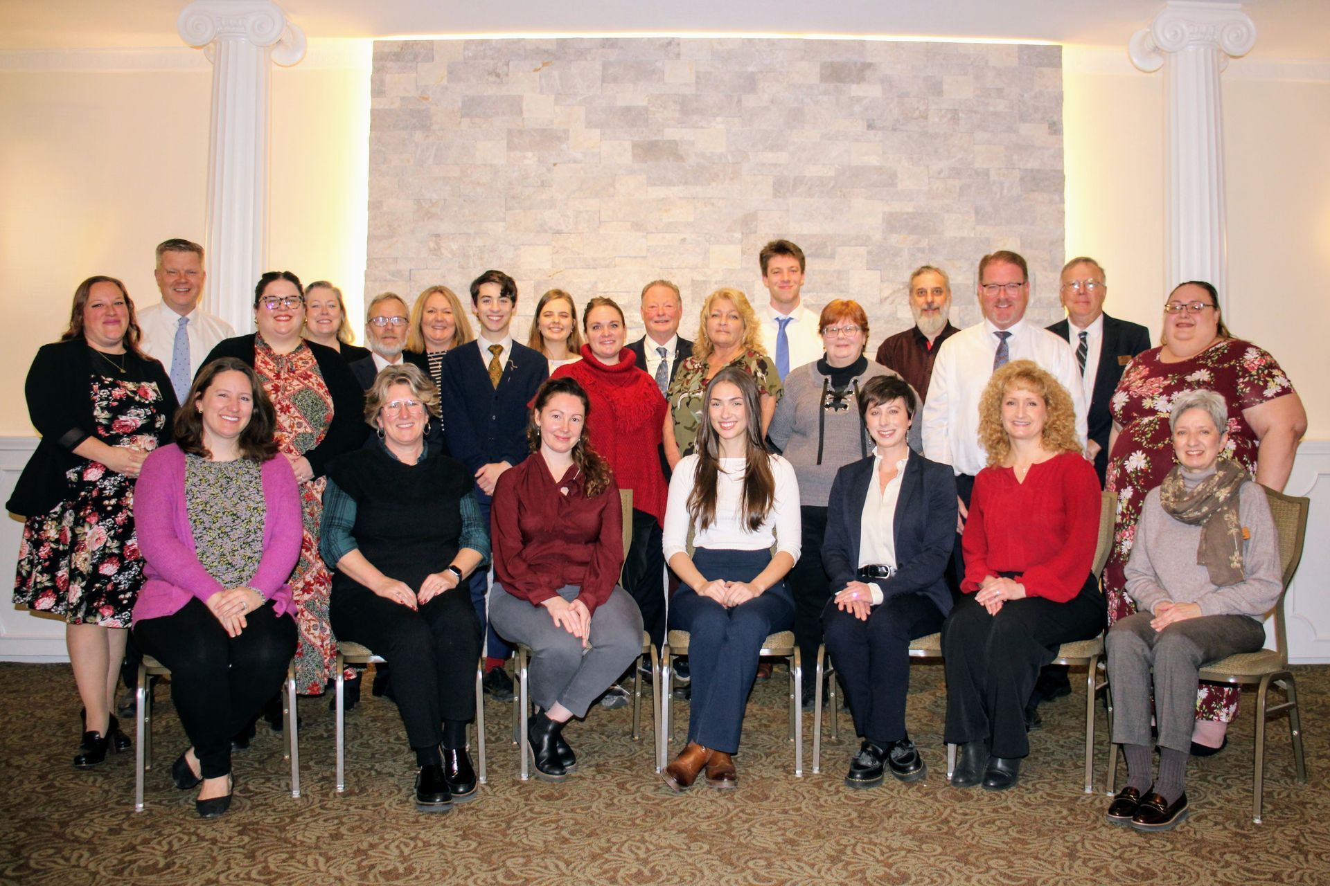 Group of people posing for a photo in a room with a stone wall backdrop; most are smiling.