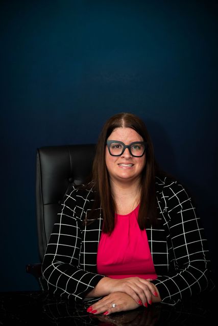 Person seated in a black chair, smiling, wearing glasses, a pink top, and a black checkered blazer against a dark background