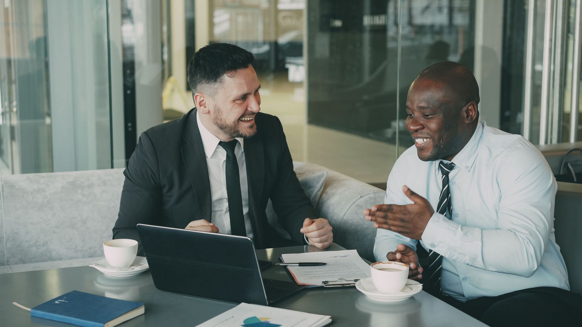 Two coworkers in business attire smiling and talking at a desk with a laptop and coffee cups