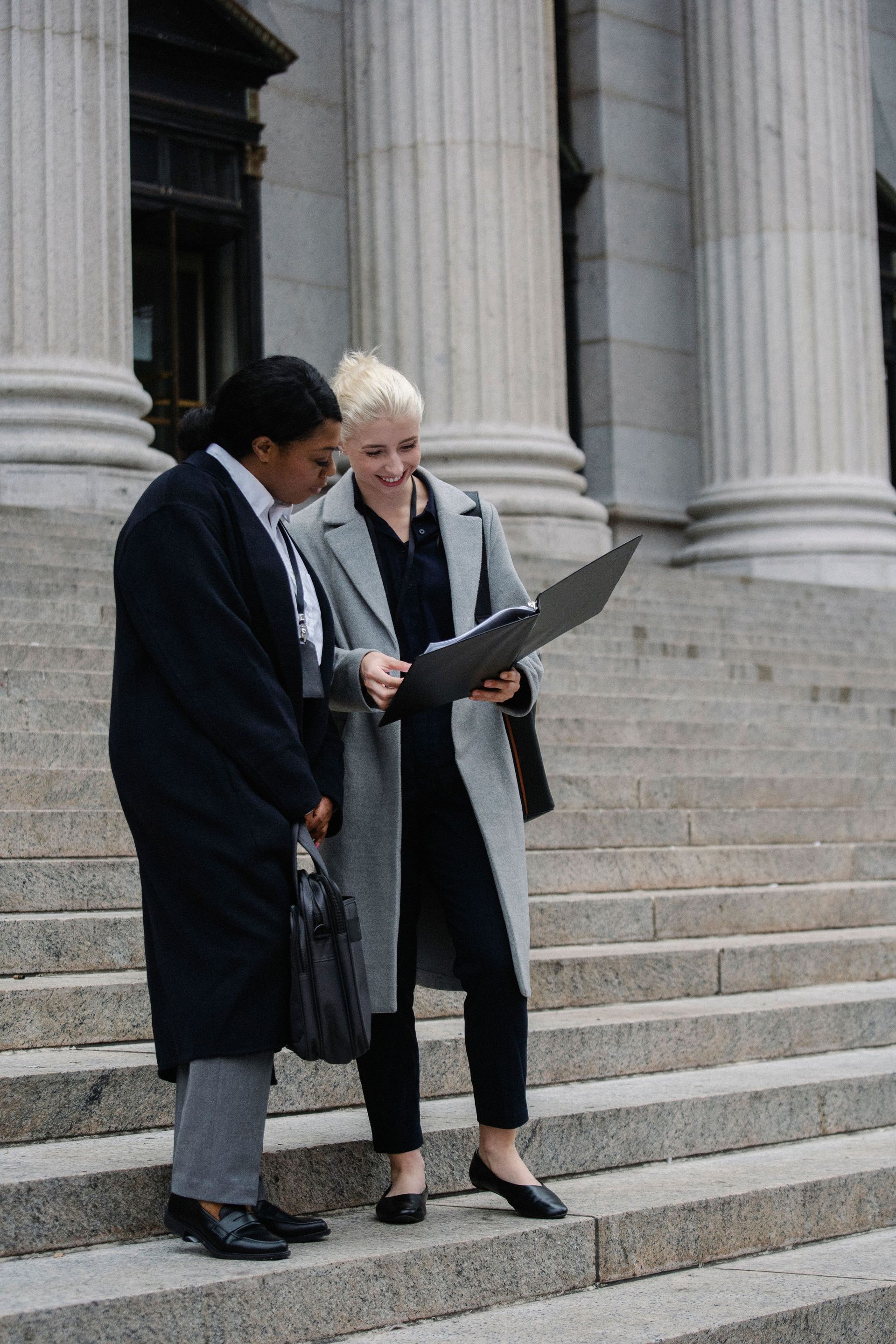 Two people in dark coats review a folder while standing on stone steps outside a large building.