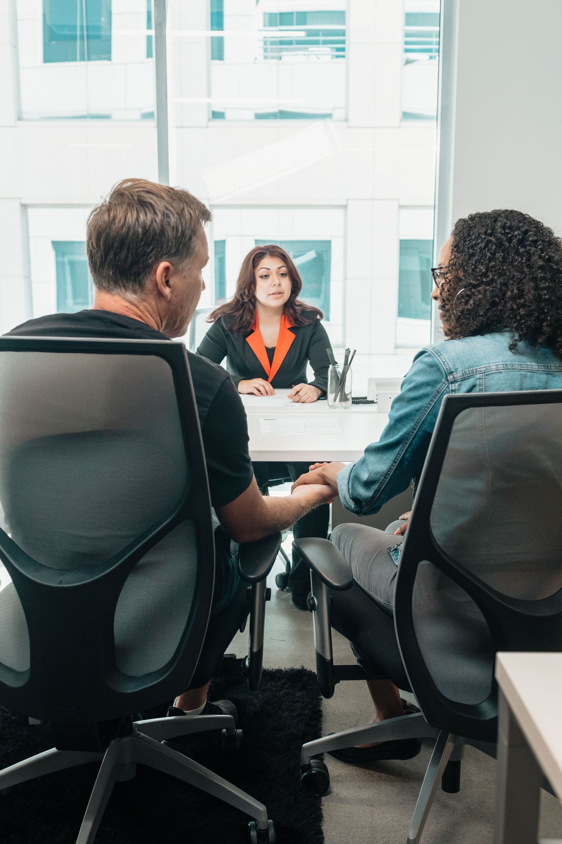 Meeting in a modern office with three people seated around a table, facing a woman speaking across from them.