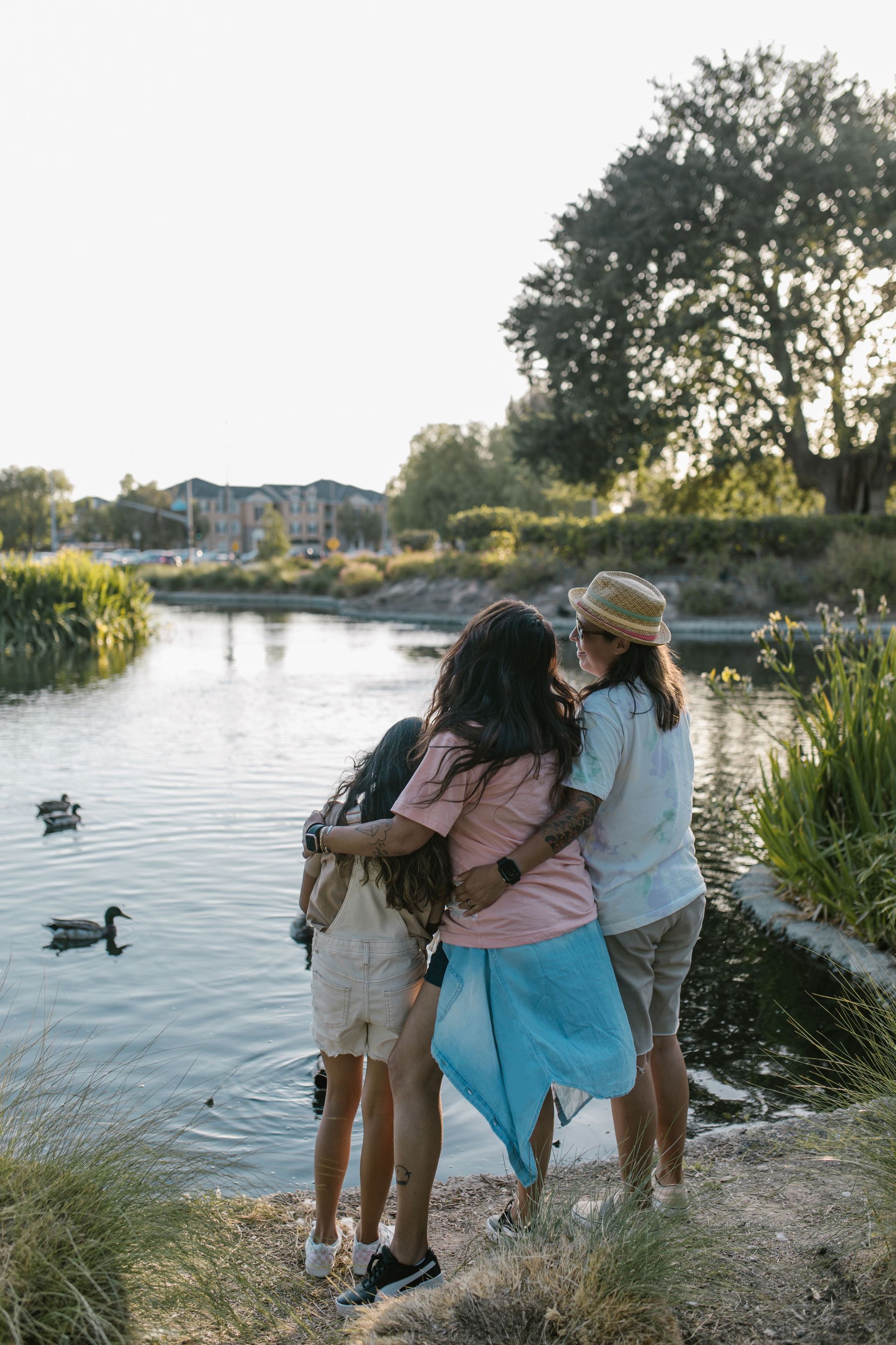 Three people hugging by a lakeside at sunset, with ducks on the water.