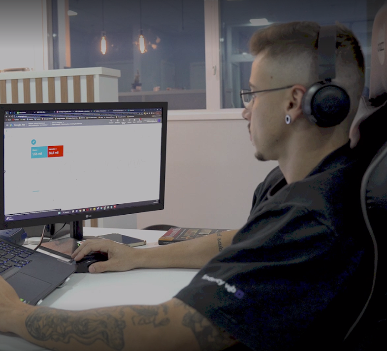 Homem com fones de ouvido trabalhando em um computador, ambiente de escritório, tatuagem no braço.