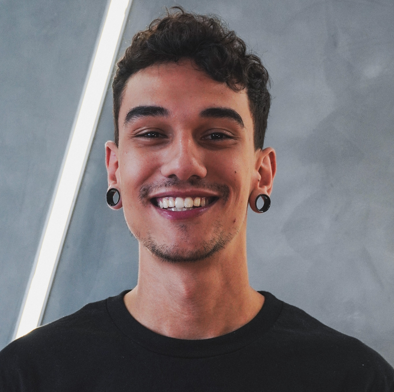 Jovem com alargadores de ouvido sorri largamente contra uma parede moderna.