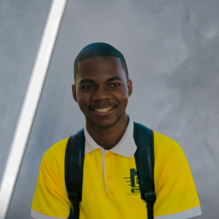Homem de camisa amarela sorrindo, usando uma mochila, em frente a uma parede cinza e branca.