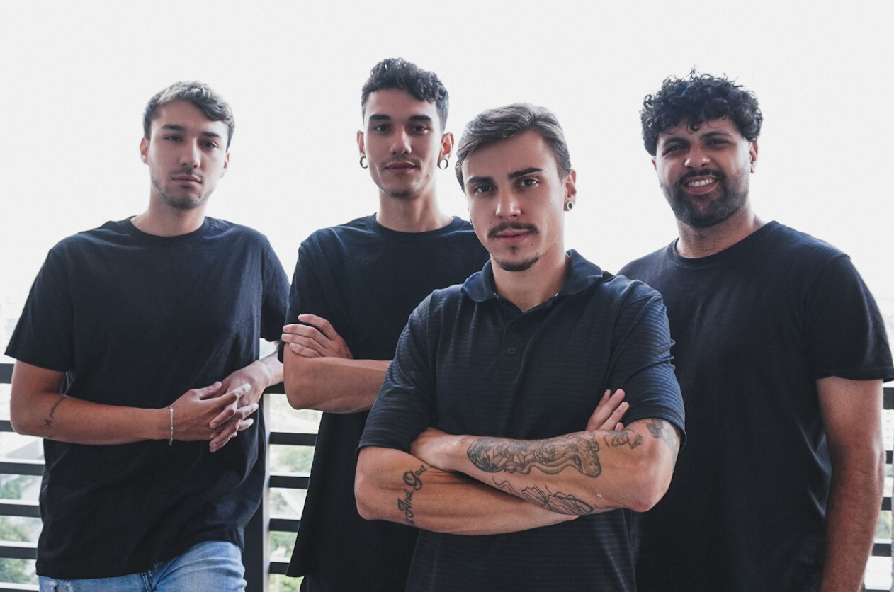 Quatro homens de camisas pretas, braços cruzados ou ao lado do corpo, em pé do lado de fora, com um fundo claro.