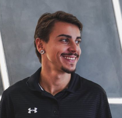 Jovem com barba rala e expressão sorridente. Vestindo uma camisa polo preta, fundo cinza.