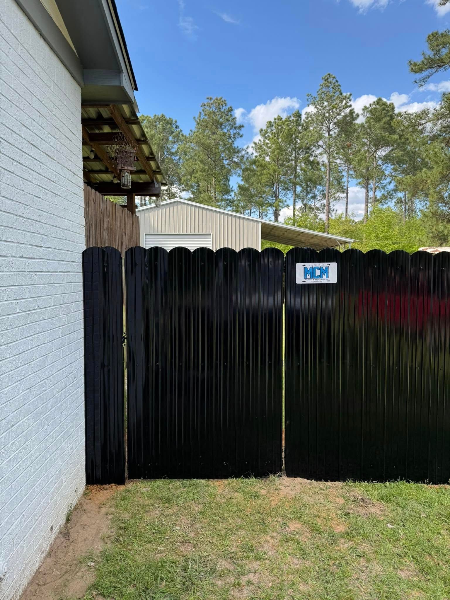 Black fence beside a white house with a small NCN sign, grassy yard, and trees under a blue sky