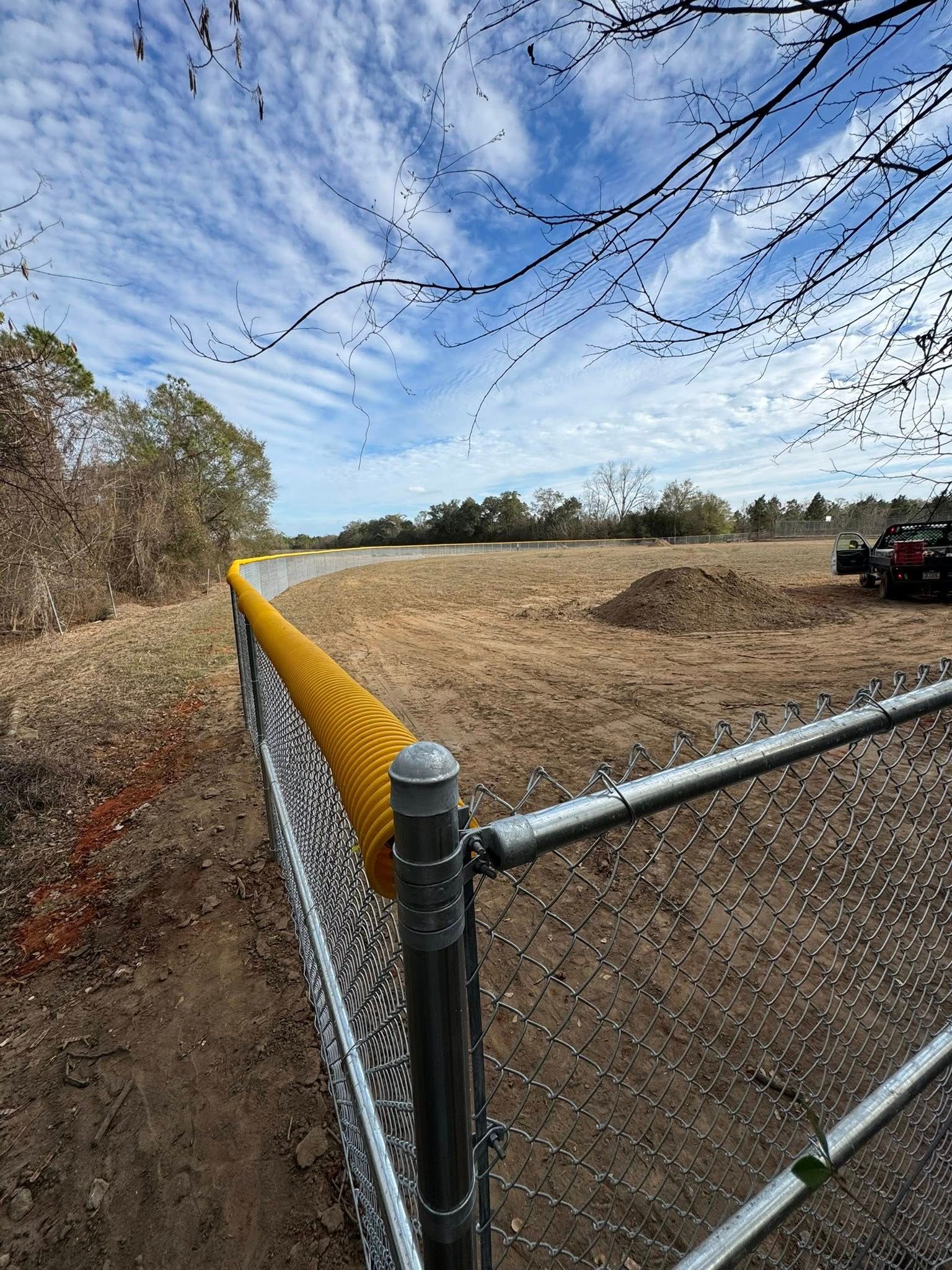 Wide farm field beside a chain-link fence under a blue sky, with a utility vehicle in the distance.