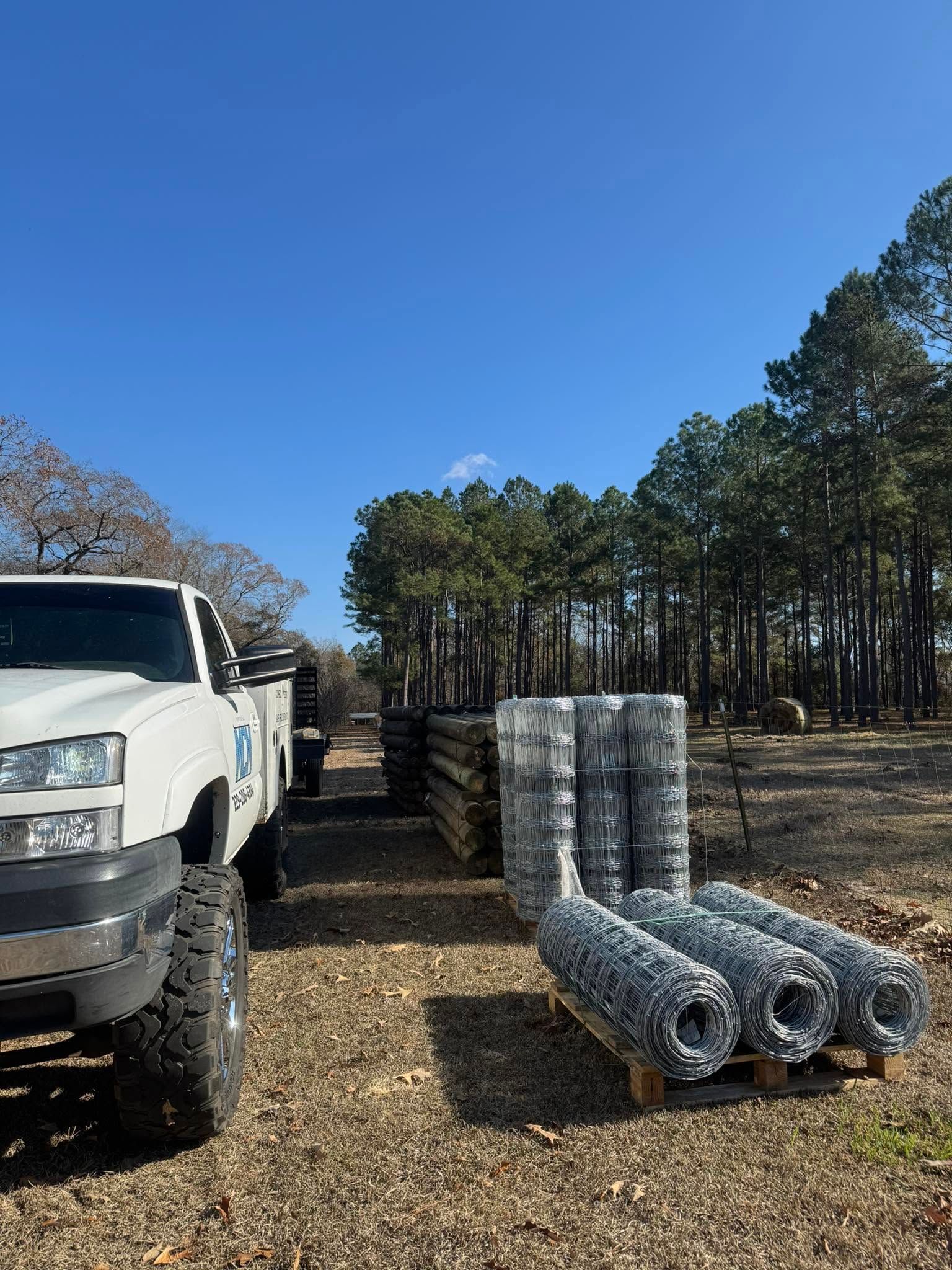 White truck beside rolls of wire mesh on a gravel lot with trees and blue sky