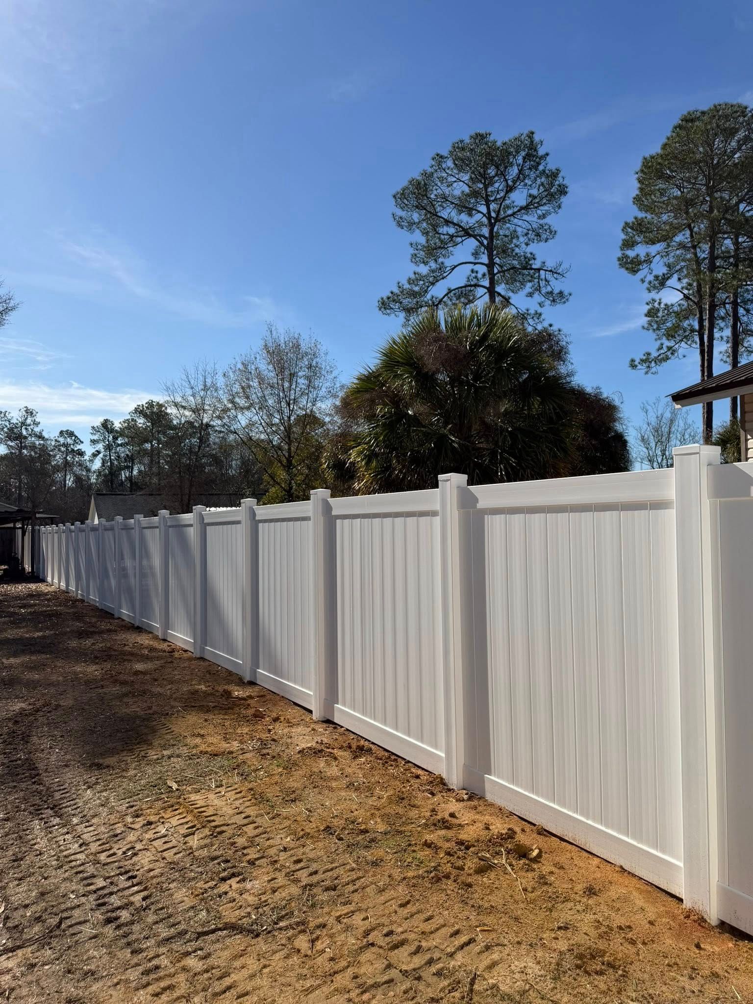 White vinyl fence along a dirt path under a clear blue sky with trees in the background