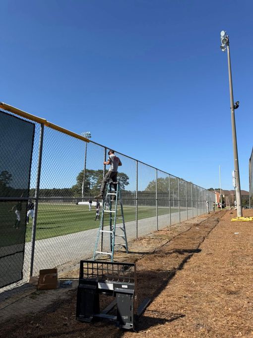 Person on a ladder fixing a chain-link fence beside a sports field under a clear blue sky