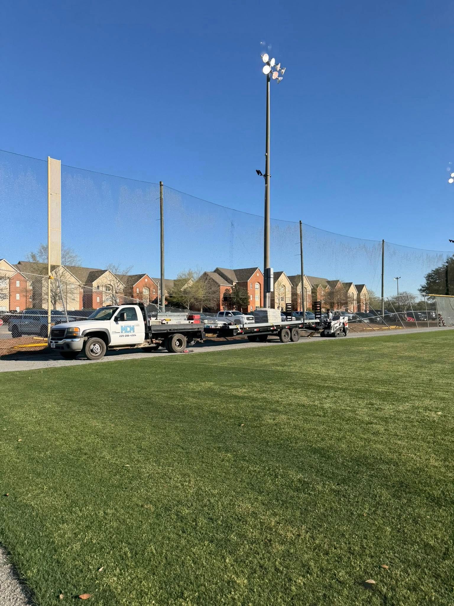 Baseball field with green grass, parked trucks along the fence, and a clear blue sky
