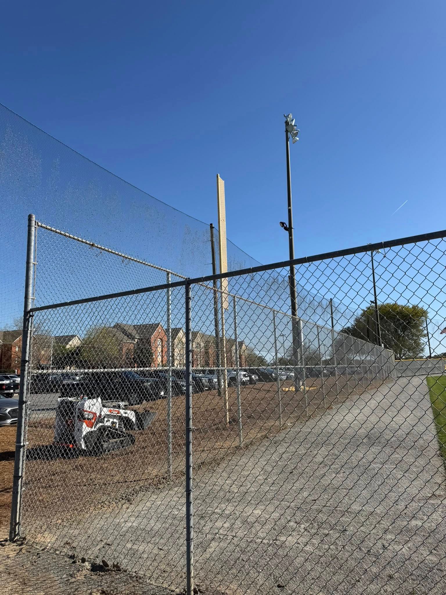 Baseball field behind chain-link fence under a clear blue sky, with a parked car and light poles.