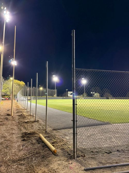 Lit sports field at night behind a chain-link fence, with tall floodlights and gravel in the foreground