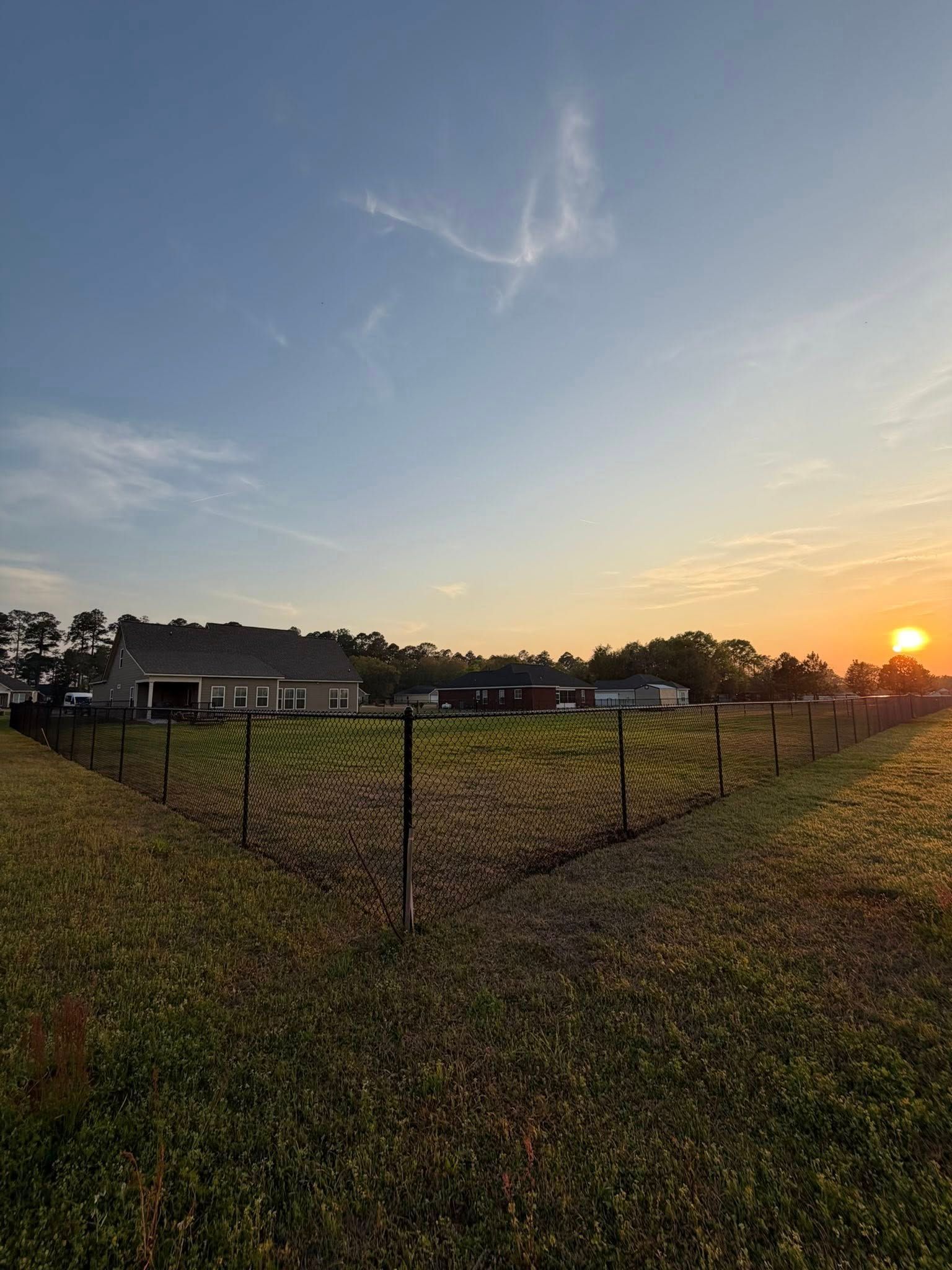 Sunset over a fenced grassy field with buildings and trees in the distance