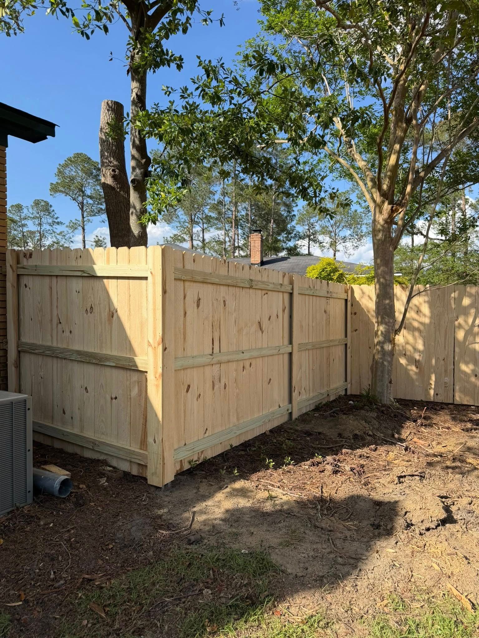 Wooden fenced enclosure in a backyard with trees and dirt ground in sunlight