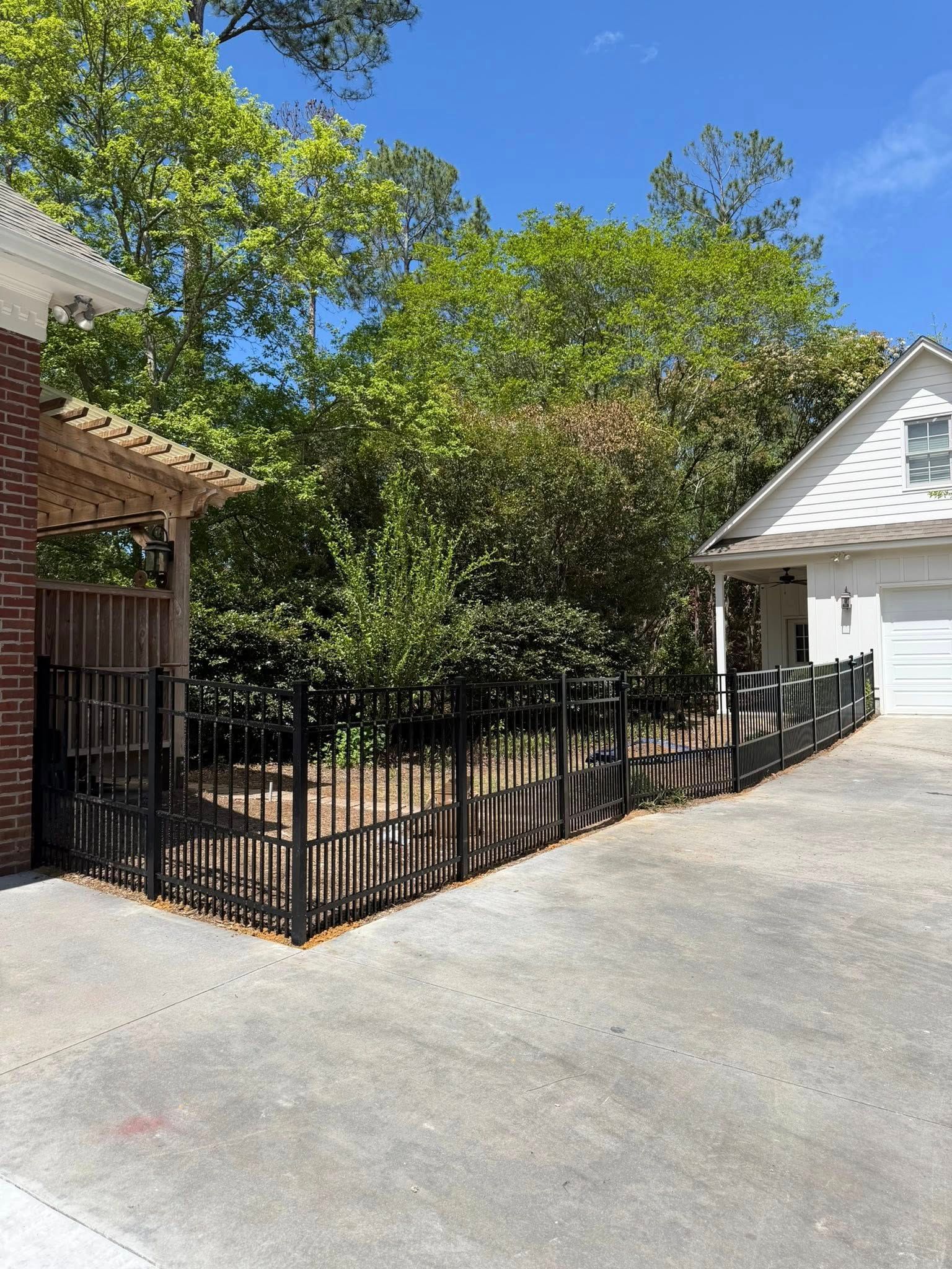Black metal fence around a leafy courtyard beside a concrete driveway and white house.