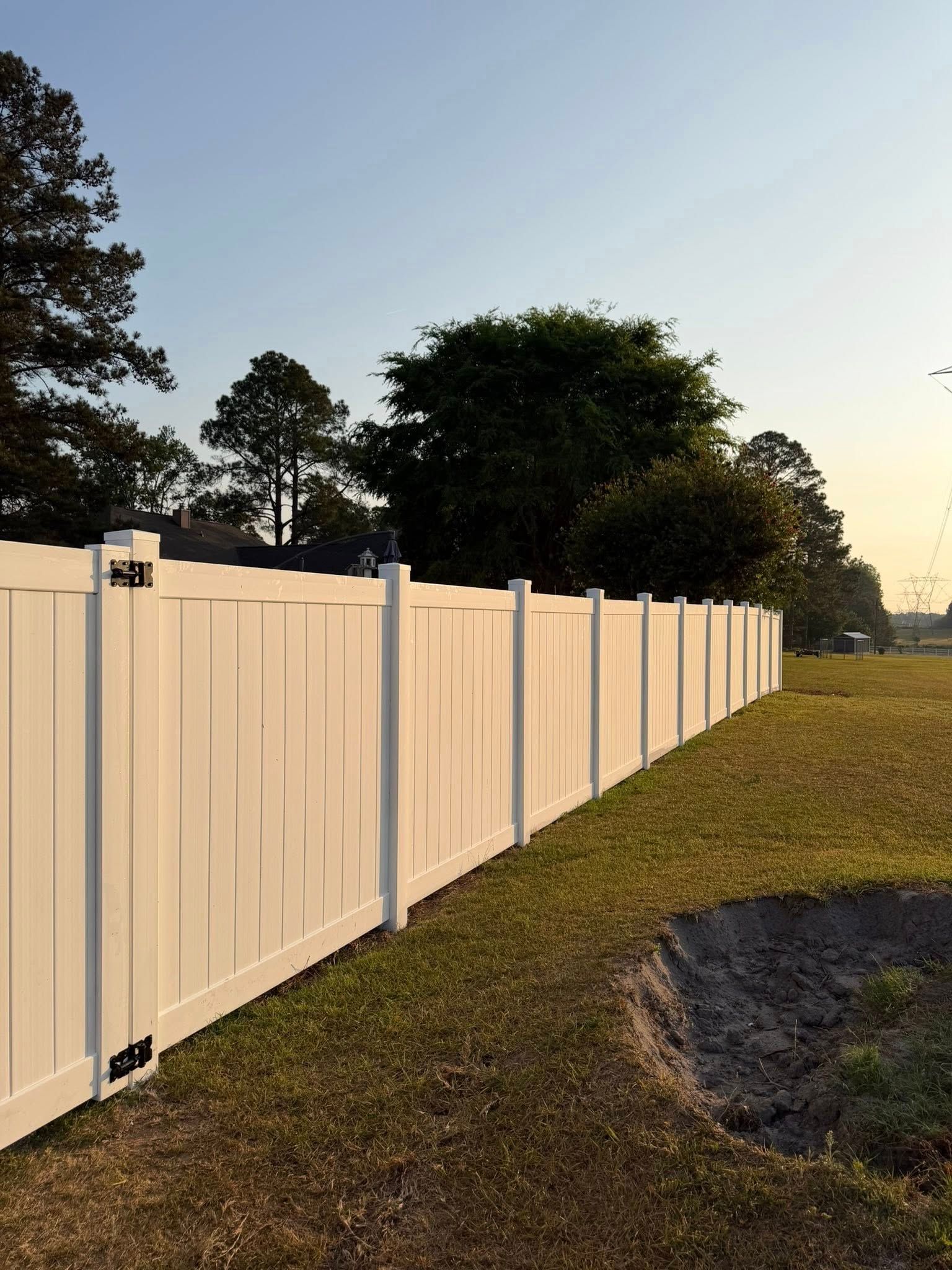 White vinyl fence along a grassy field at sunset, with trees and a clear sky.