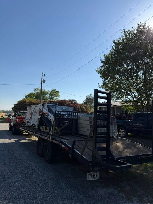 Flatbed tow truck parked on gravel road with another truck and trees under a clear blue sky