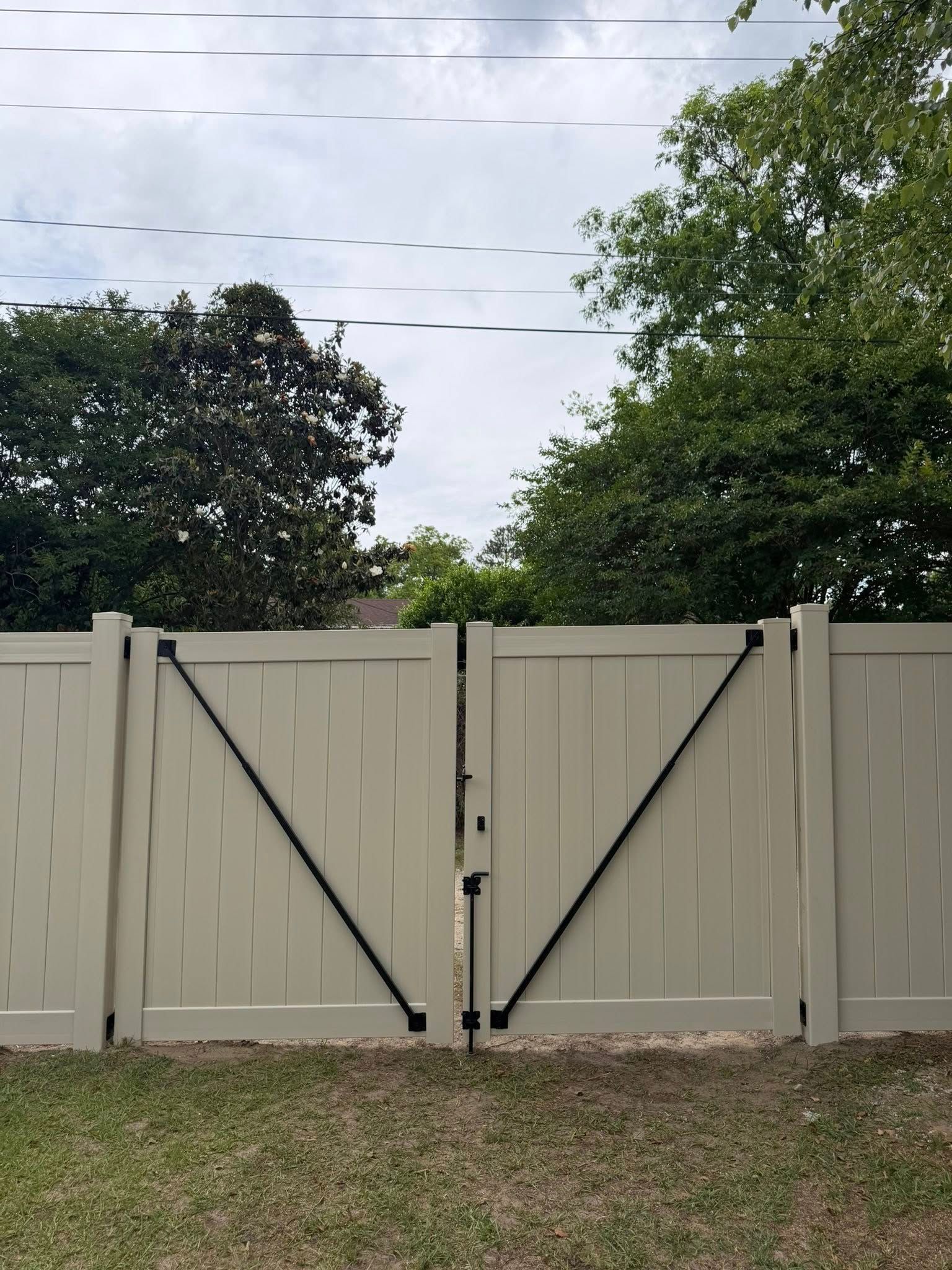 Closed beige wooden gate with black diagonal braces, between fence panels, with trees and cloudy sky behind