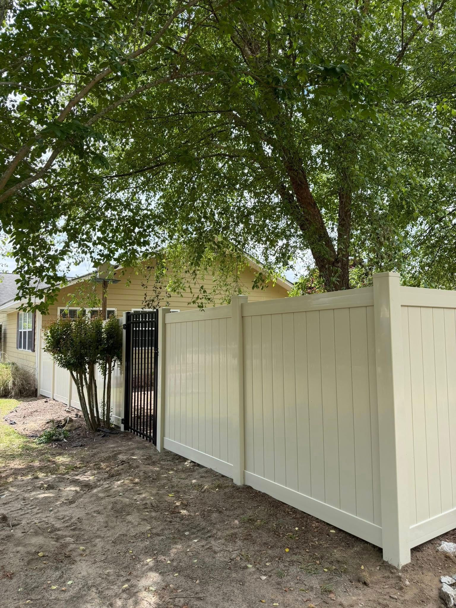 White privacy fence beside a house under a large leafy tree, with a black gate on the left.