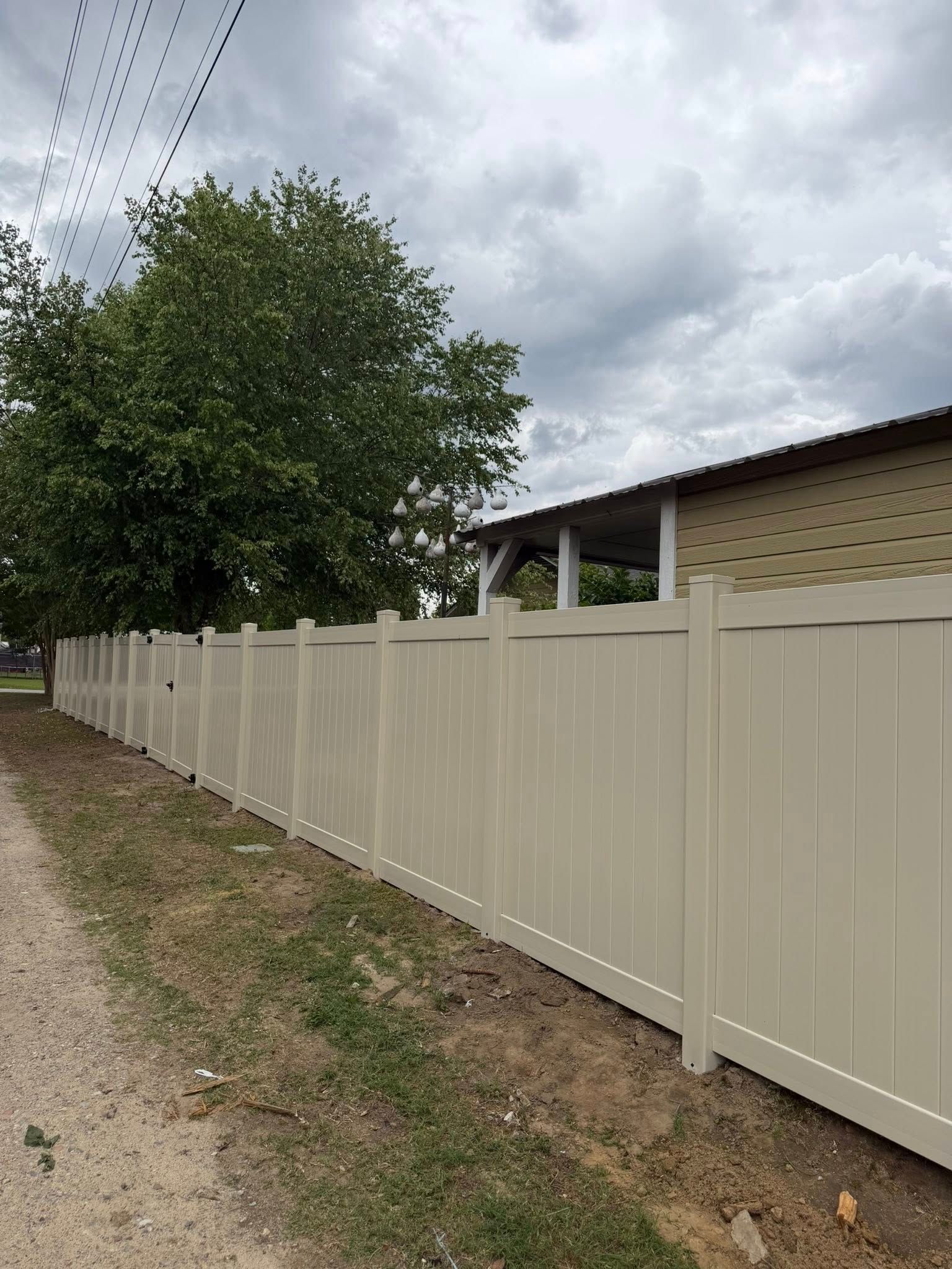 Beige vinyl fence along a dirt road beside a tree and a house under cloudy sky