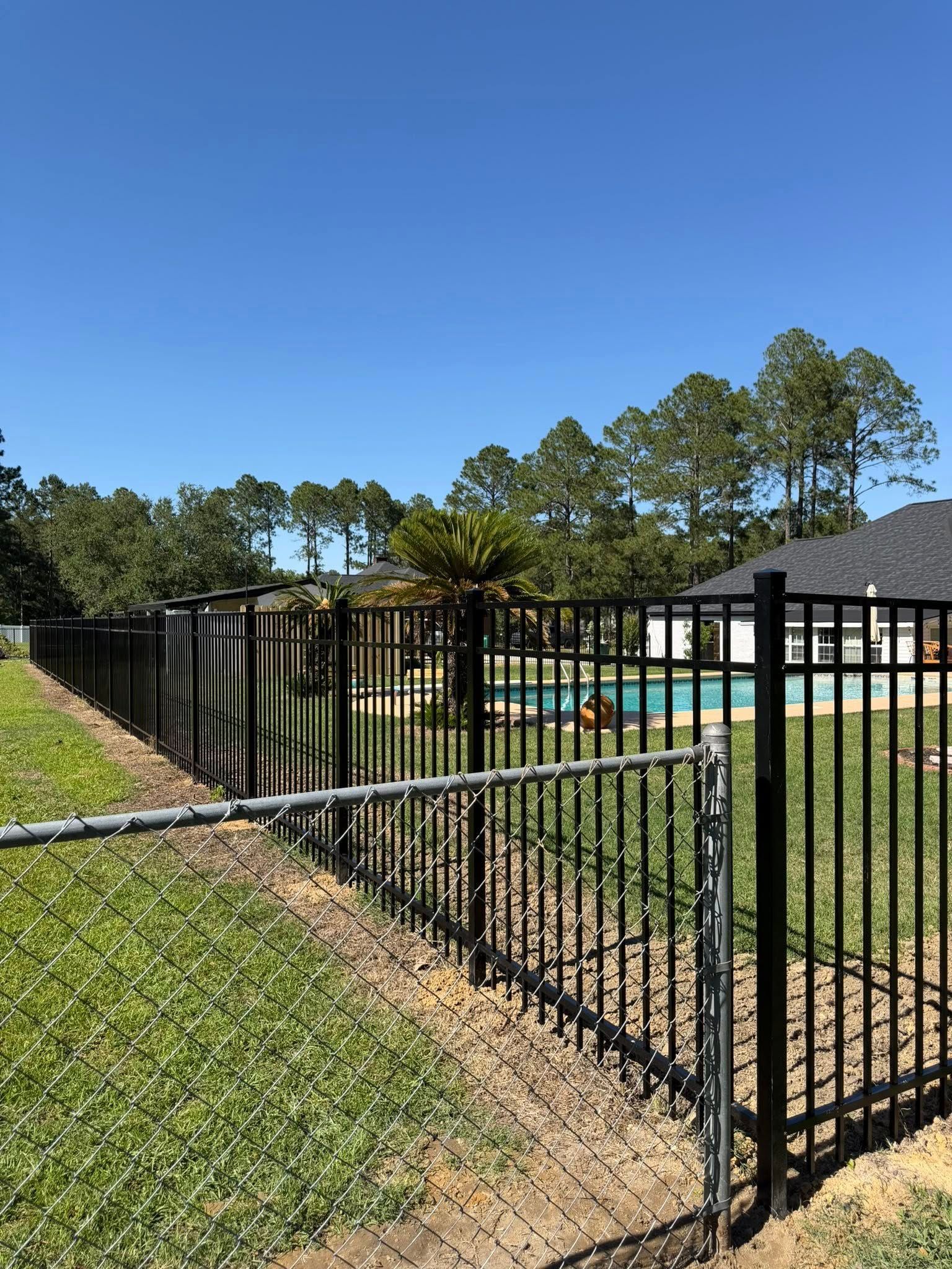 Black metal fence around a backyard pool with a house and tall trees under a clear blue sky.
