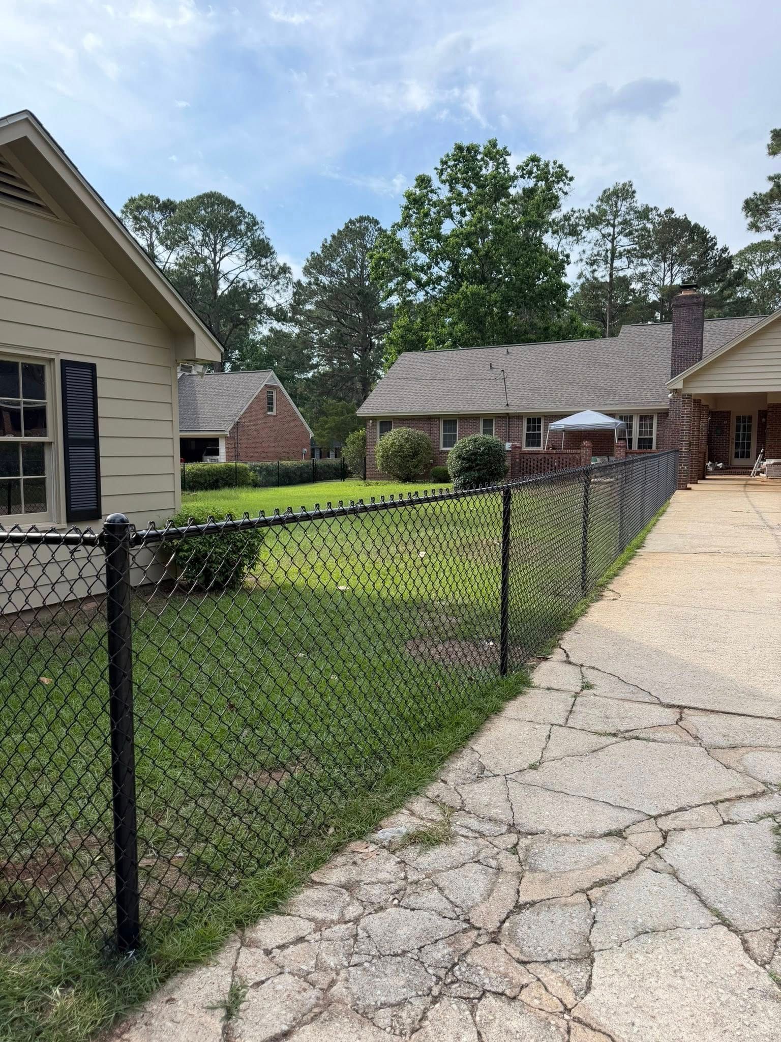 Chain-link fence along a cracked driveway beside suburban houses and a green yard.