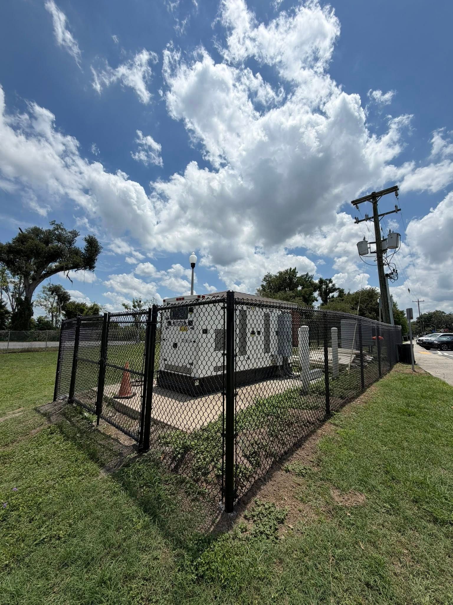 Fenced utility equipment beside a road under a bright cloudy sky