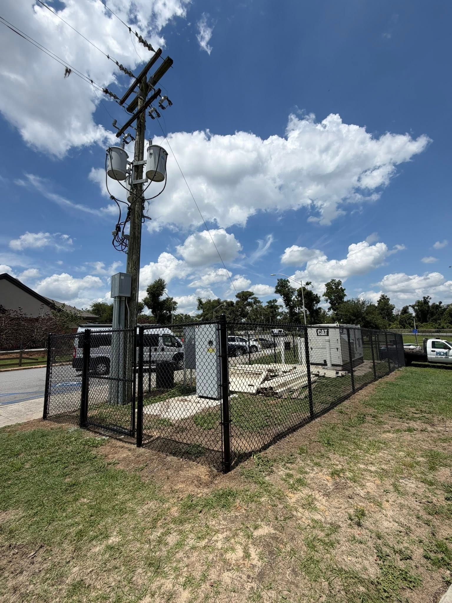 Fenced utility equipment under a utility pole beside a grassy roadside under a blue sky with clouds