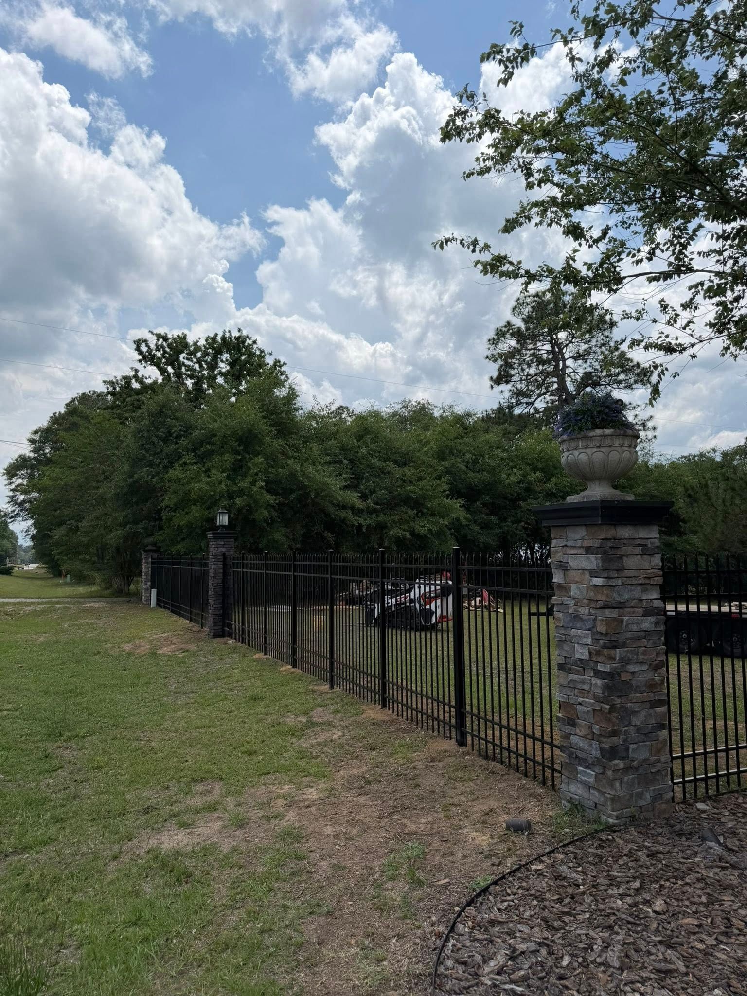 Grassy path beside a black iron fence and stone pillars under a partly cloudy sky