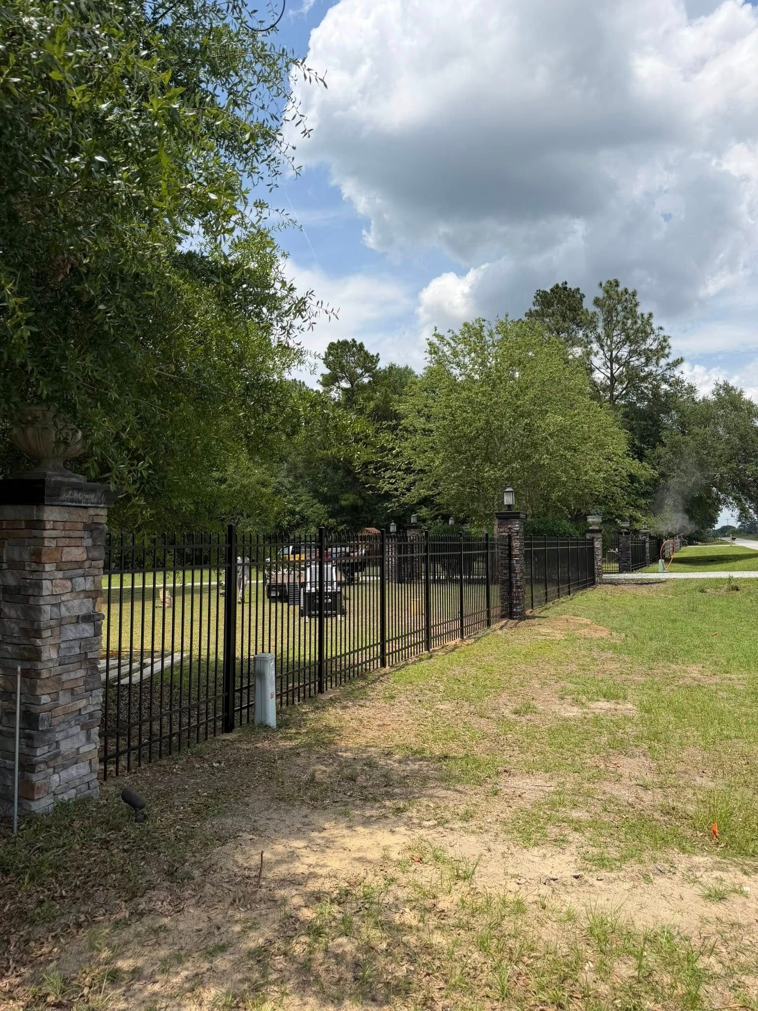 Black metal fence beside a grassy roadside under cloudy skies, with trees and a stone pillar