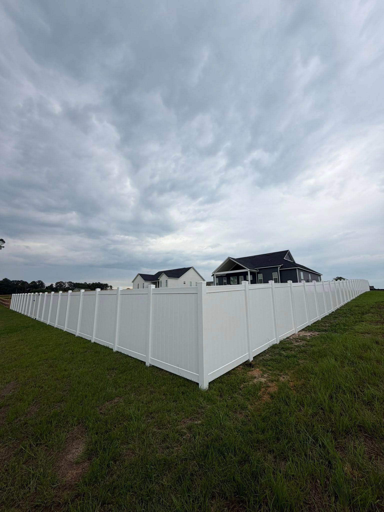 White vinyl fence enclosing a grassy yard under a cloudy sky, with houses in the background.