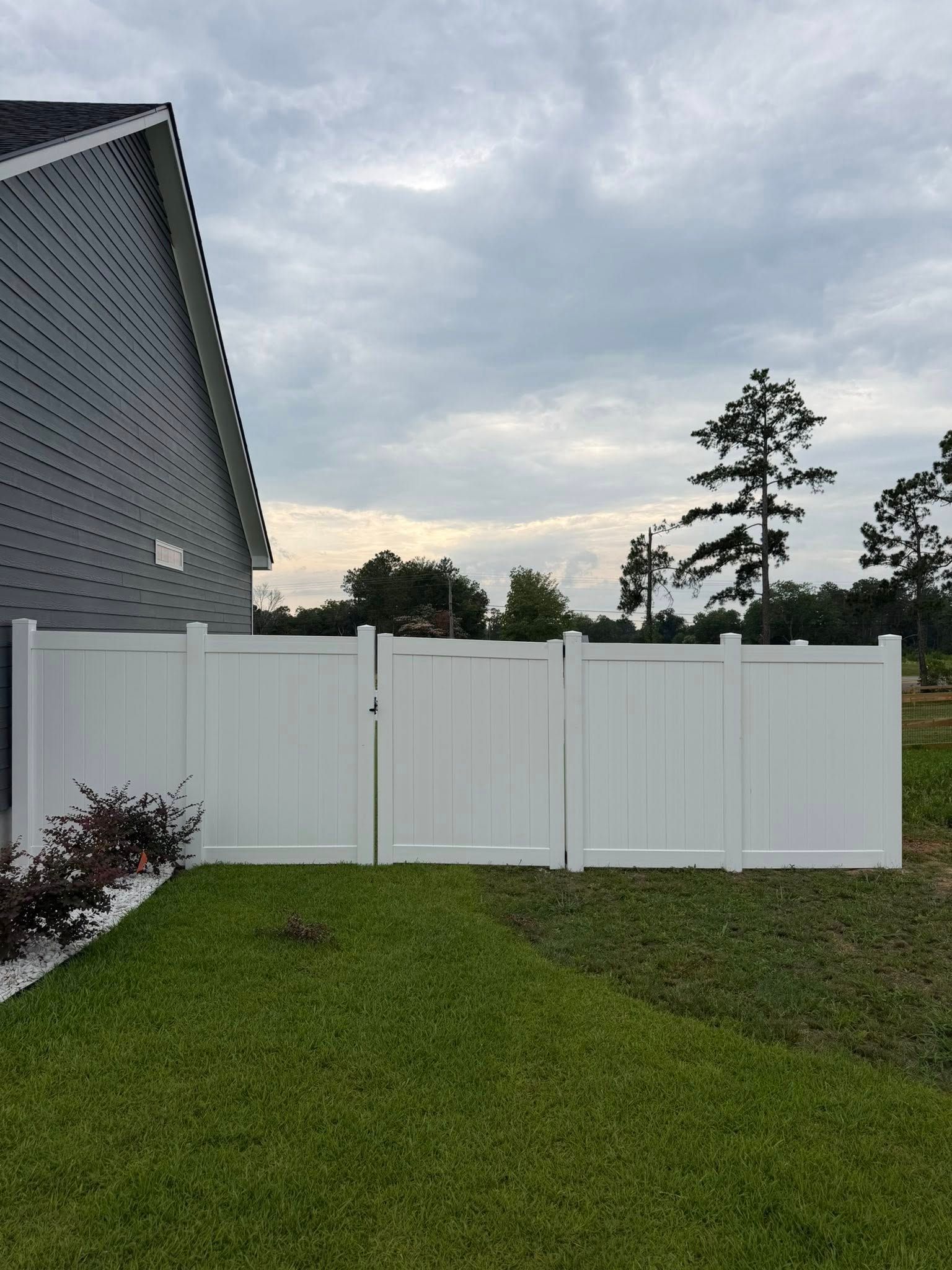 White privacy fence in a green backyard beside a gray house under an overcast sky