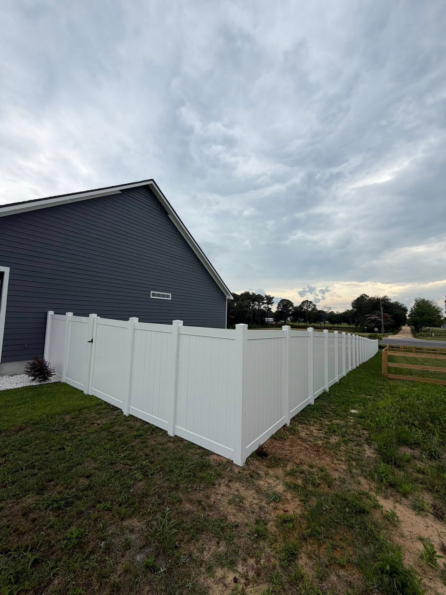 White vinyl privacy fence beside a dark gray house under a cloudy sky, with grassy yard and open field beyond
