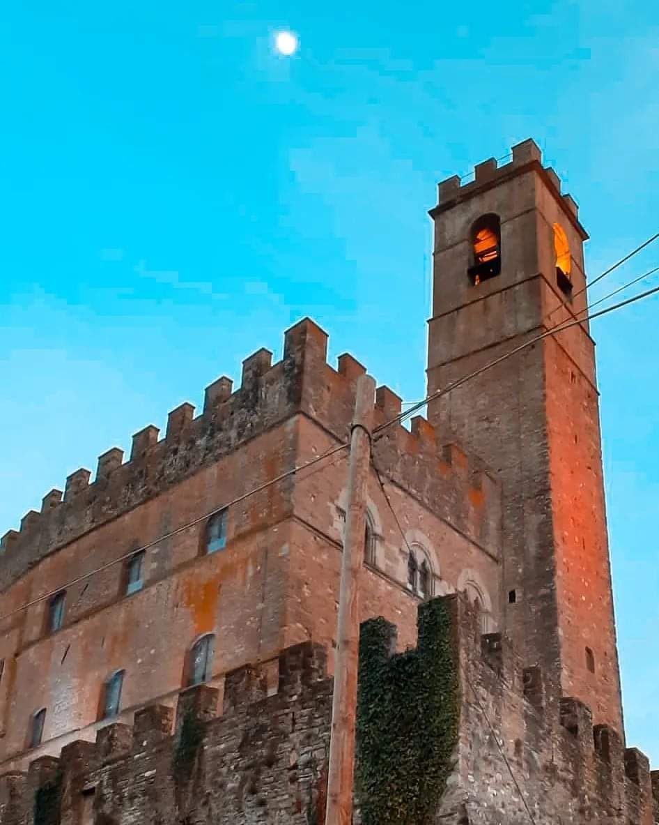 Torre di pietra del castello con merlature al crepuscolo contro un cielo azzurro, una finestra che brilla di un arancione intenso.