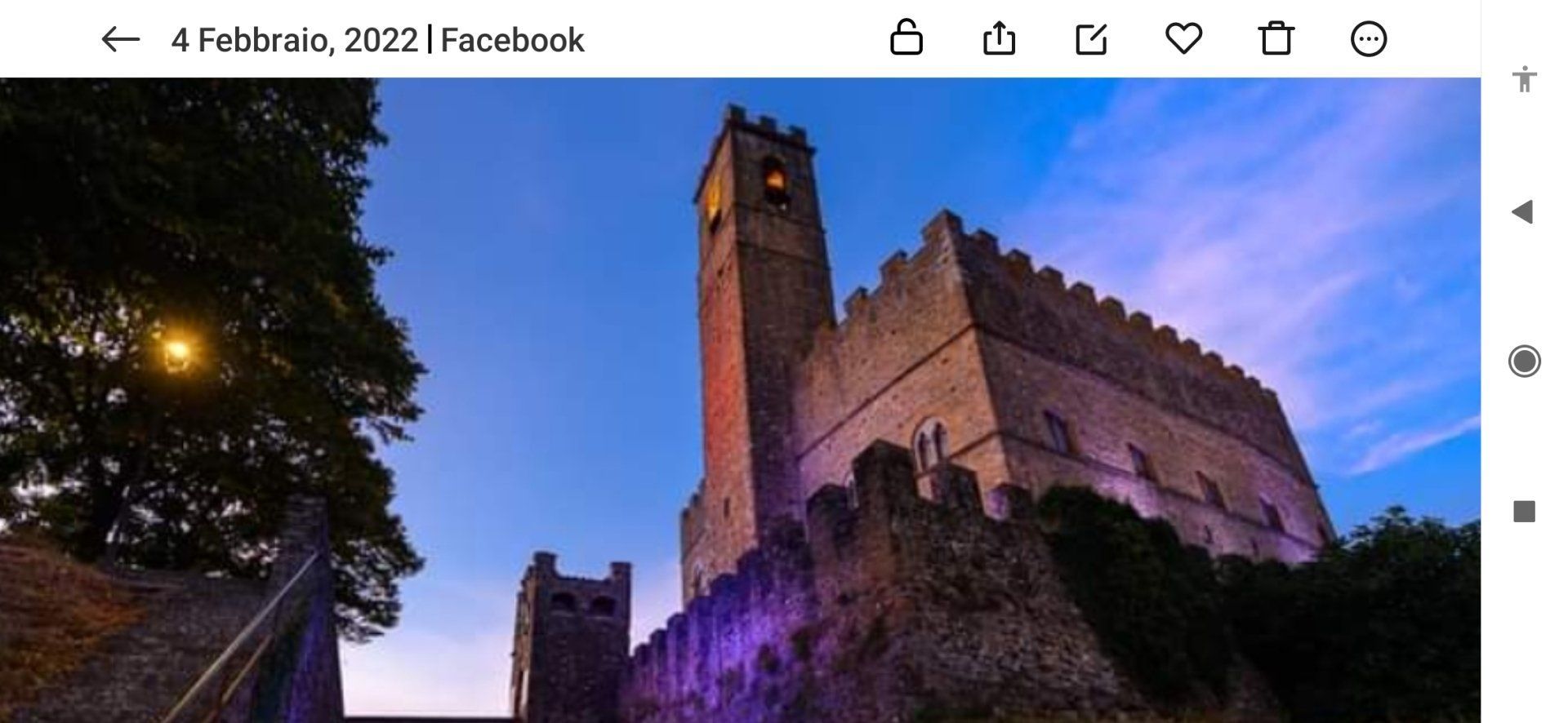 Torre di pietra del castello illuminata di viola al crepuscolo, sotto un cielo crepuscolare, con alberi in primo piano.