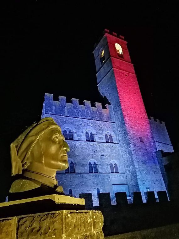Torre del castello illuminata di notte con luci rosse e blu, vista da dietro una statua a busto dorato.