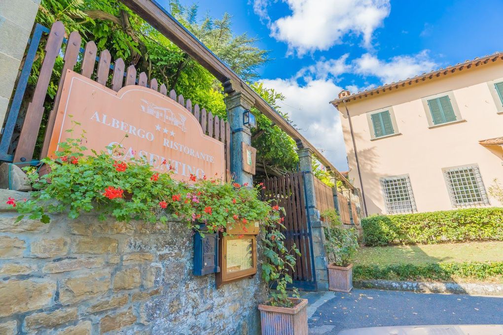 Cancello in pietra con insegna rosa, fiori e rampicanti verdi accanto a un edificio color crema sotto un cielo azzurro.